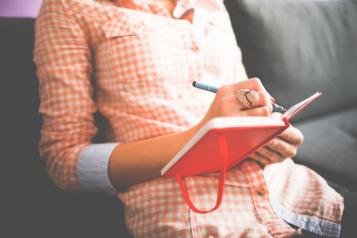 Woman in checked shirt sitting down and writing in a red notebook