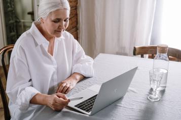Grey-haired woman in white shirt sits at table with laptop
